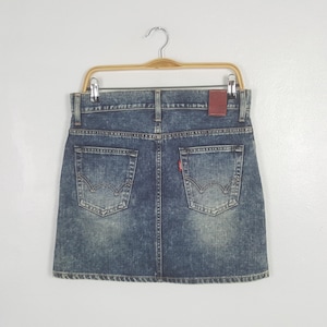 Puede incluir: Minifalda vaquera en un lavado azul claro, colgada de una percha de madera. La falda presenta dos bolsillos traseros con un dise&ntilde;o distintivo, una etiqueta roja y un parche de cuero marr&oacute;n. La falda es de mezclilla.