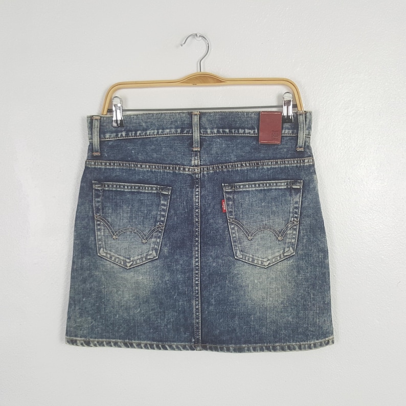 Puede incluir: Minifalda vaquera en un lavado azul claro, colgada de una percha de madera. La falda presenta dos bolsillos traseros con un dise&ntilde;o distintivo, una etiqueta roja y un parche de cuero marr&oacute;n. La falda es de mezclilla.