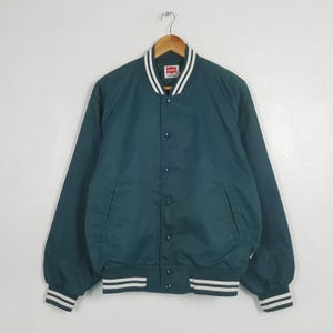 以下が含まれることがあります： 襟、袖口、裾に白と黒のストライプのアクセントが付いた、ティール色のボンバージャケット。 ジャケットはボタン留め、フロントポケット2つ、襟にブランドラベルが付いています。 木製ハンガーに吊るされています。