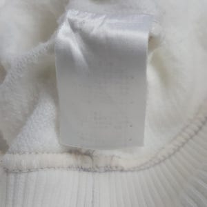 May include: A white fabric garment featuring a rectangular white care label sewn onto the interior. The label is positioned vertically against the soft, textured white fleece material of the clothing item, which includes a ribbed hem detail.