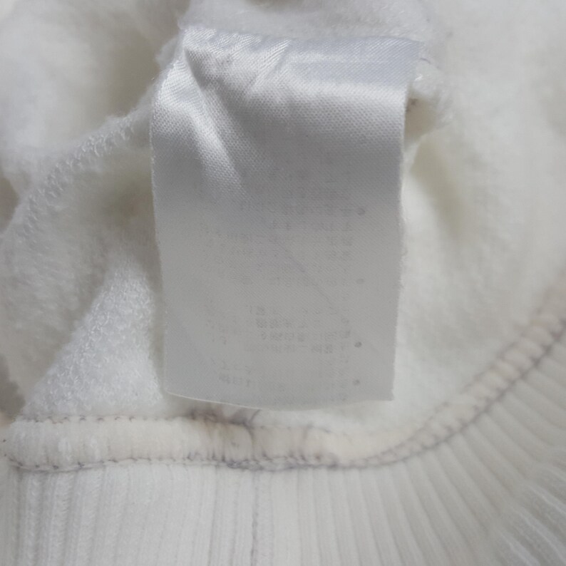 May include: A white fabric garment featuring a rectangular white care label sewn onto the interior. The label is positioned vertically against the soft, textured white fleece material of the clothing item, which includes a ribbed hem detail.