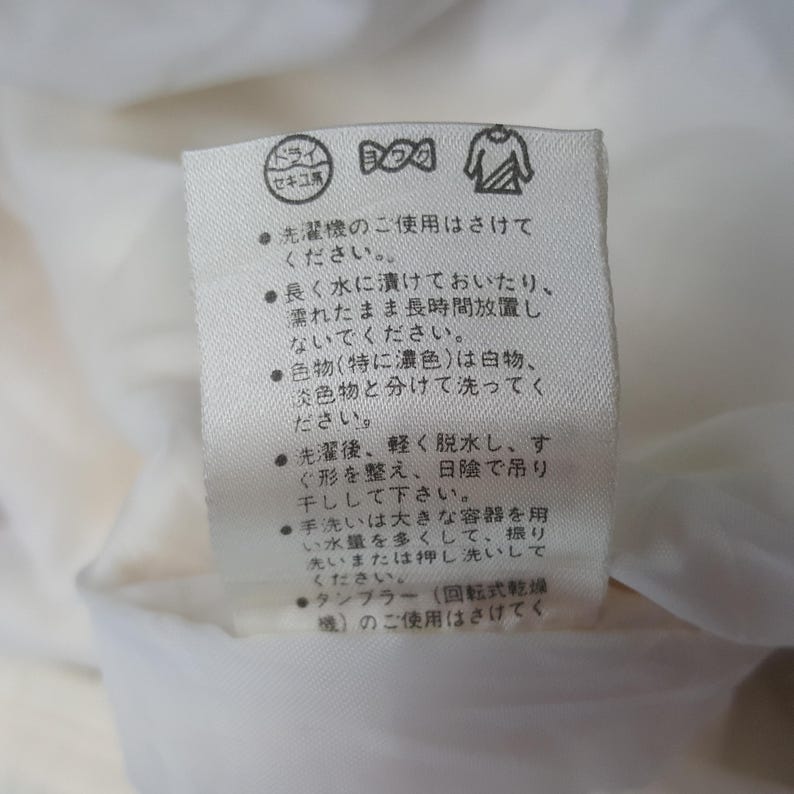 Pode incluir: Uma etiqueta de tecido branco com texto japon&ecirc;s e s&iacute;mbolos de cuidados. A etiqueta inclui s&iacute;mbolos para limpeza a seco, lavagem e secagem. O texto fornece orienta&ccedil;&otilde;es de lavagem e secagem.