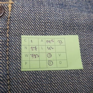 May include: Close-up of blue denim fabric with a visible weave pattern. A small green tag with black text and numbers is attached to the fabric. The tag includes the numbers 1, 31, 37, 42, and 722.