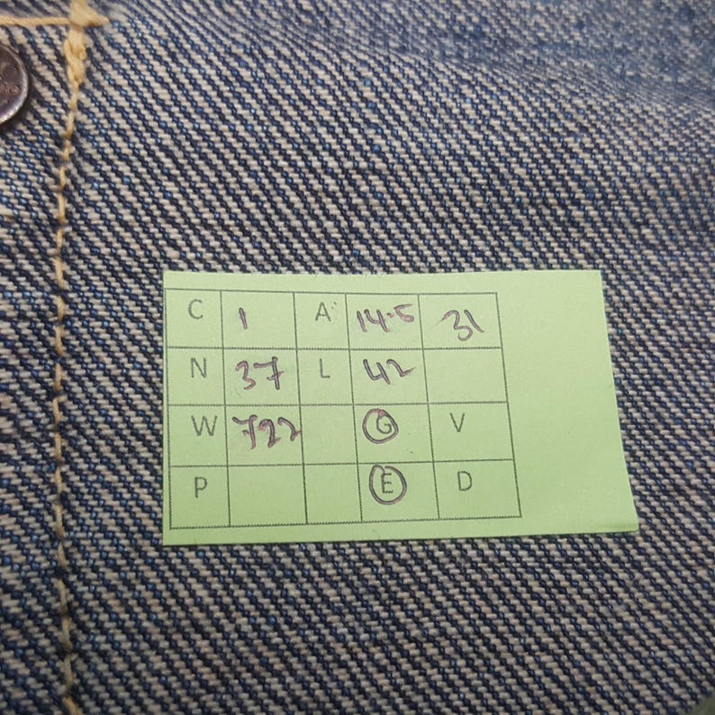 May include: Close-up of blue denim fabric with a visible weave pattern. A small green tag with black text and numbers is attached to the fabric. The tag includes the numbers 1, 31, 37, 42, and 722.