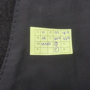 May include: Close-up of a dark gray fabric with a yellow sticky note attached. The note contains handwritten measurements and letters. The fabric has a subtle texture and appears to be part of a garment.