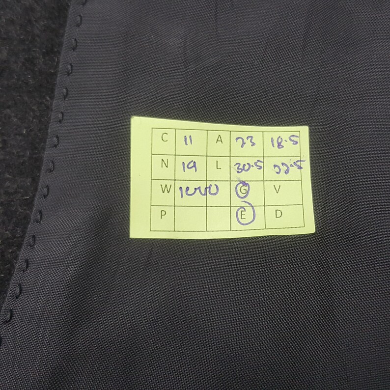 May include: Close-up of a dark gray fabric with a yellow sticky note attached. The note contains handwritten measurements and letters. The fabric has a subtle texture and appears to be part of a garment.