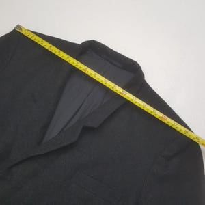 May include: A dark gray blazer with a two-button closure and a notched lapel. A yellow measuring tape is laid across the blazer, showing a measurement of approximately 19 inches. The blazer has a pocket on the left side.