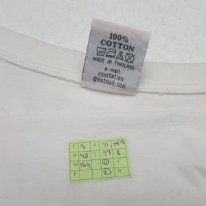 Puede incluir: Una camiseta de algod&oacute;n blanca con una etiqueta que dice "100% Algod&oacute;n" y "Made in Thailand". La etiqueta tambi&eacute;n incluye una direcci&oacute;n de correo electr&oacute;nico. Tambi&eacute;n es visible una peque&ntilde;a pegatina verde con n&uacute;meros y letras manuscritos.