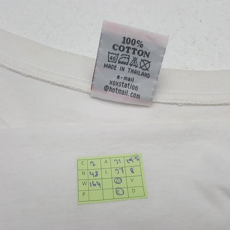 Puede incluir: Una camiseta de algod&oacute;n blanca con una etiqueta que dice "100% Algod&oacute;n" y "Made in Thailand". La etiqueta tambi&eacute;n incluye una direcci&oacute;n de correo electr&oacute;nico. Tambi&eacute;n es visible una peque&ntilde;a pegatina verde con n&uacute;meros y letras manuscritos.