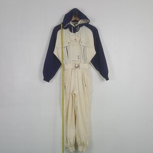 Pode incluir: Um fato de esqui vintage em creme, azul marinho e bege. O fato de uma pe&ccedil;a apresenta um design com capuz, cintura com cinto e v&aacute;rios bolsos. As mangas e o capuz s&atilde;o azul marinho, enquanto o corpo &eacute; creme. Fecho de correr e material resistente &agrave; &aacute;gua.