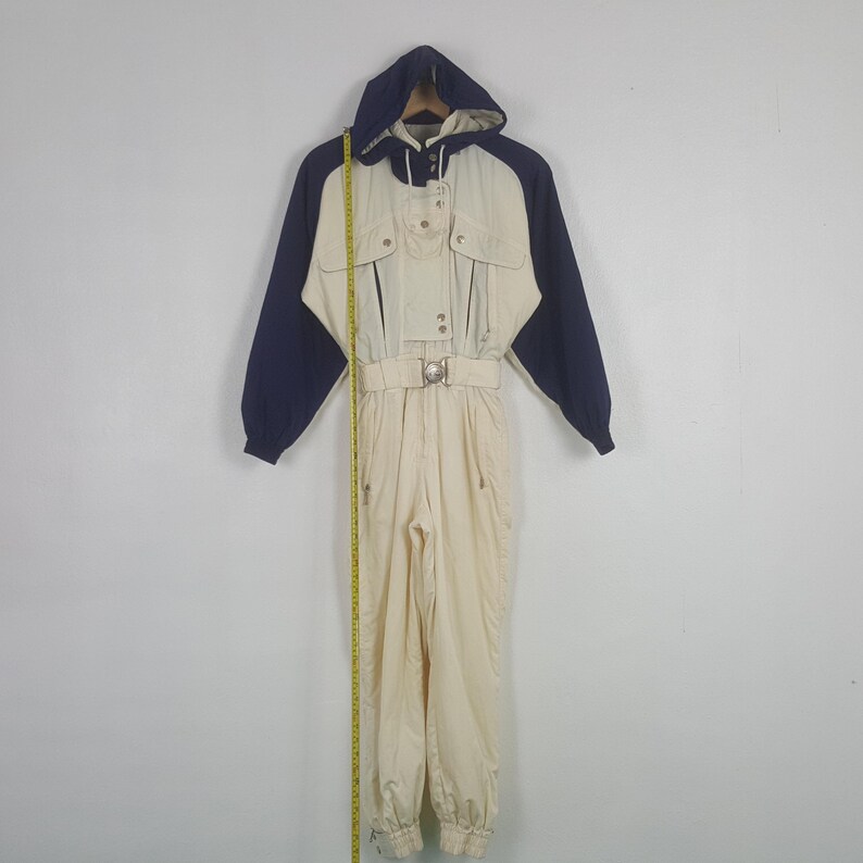 Pode incluir: Um fato de esqui vintage em creme, azul marinho e bege. O fato de uma pe&ccedil;a apresenta um design com capuz, cintura com cinto e v&aacute;rios bolsos. As mangas e o capuz s&atilde;o azul marinho, enquanto o corpo &eacute; creme. Fecho de correr e material resistente &agrave; &aacute;gua.