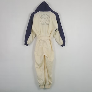 Pode incluir: Macac&atilde;o vintage creme com capuz e mangas azul marinho. A parte de tr&aacute;s apresenta o texto "sport fun" acima das letras "AC" e a palavra "courr&egrave;ges". O macac&atilde;o tem cintura ajustada e punhos el&aacute;sticos.