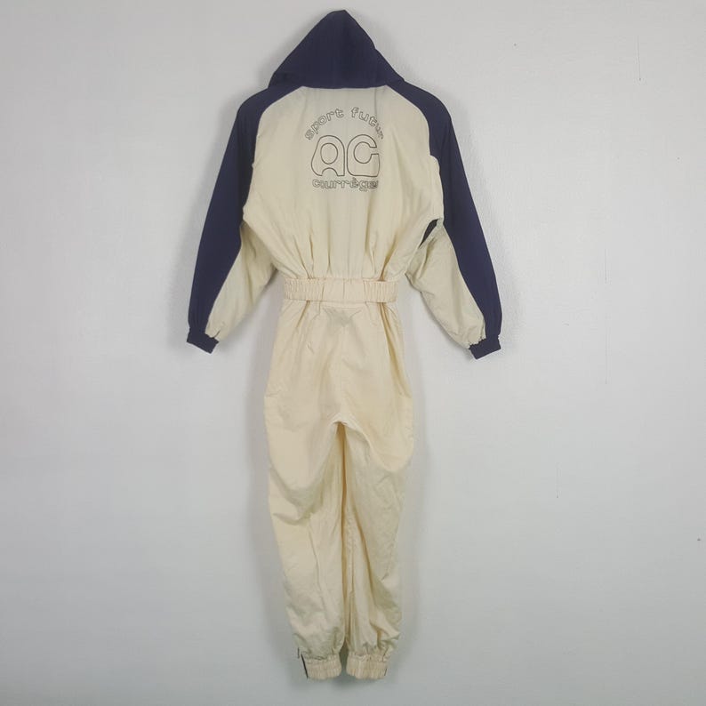 Pode incluir: Macac&atilde;o vintage creme com capuz e mangas azul marinho. A parte de tr&aacute;s apresenta o texto "sport fun" acima das letras "AC" e a palavra "courr&egrave;ges". O macac&atilde;o tem cintura ajustada e punhos el&aacute;sticos.