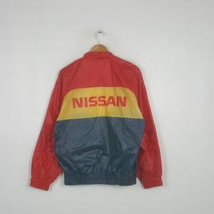 Puede incluir: Una chaqueta cortavientos de color rojo, amarillo y azul oscuro con la palabra NISSAN impresa en rojo sobre una franja amarilla horizontal. La prenda tiene mangas largas con puños elásticos, cuello alto y cintura elástica acanalada.