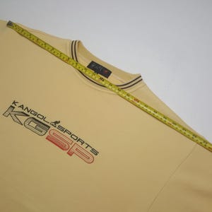 May include: A light yellow sweatshirt with the text "KANGOL SPORTS" in black and red. The collar has brown and dark brown stripes. A yellow measuring tape is across the sweatshirt.