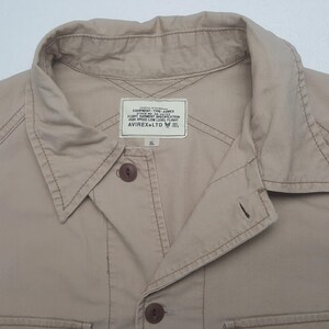 May include: Close-up of a tan button-down shirt with a collar and two visible buttons. The shirt has a label that reads "EQUIPMENT-TYPE-AVIREX FLIGHT GARMENT SPECIFICATION HIGH SPEED LOW LEVEL FLIGHT AVIREX LTD XL."