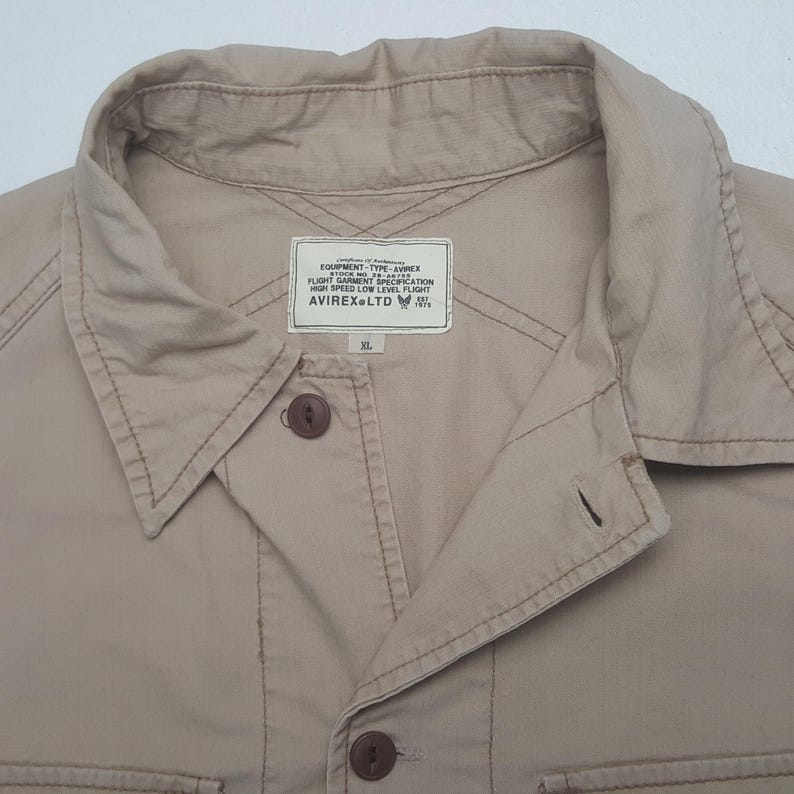 May include: Close-up of a tan button-down shirt with a collar and two visible buttons. The shirt has a label that reads "EQUIPMENT-TYPE-AVIREX FLIGHT GARMENT SPECIFICATION HIGH SPEED LOW LEVEL FLIGHT AVIREX LTD XL."