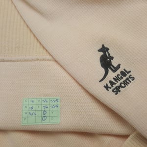 May include: A light peach-colored Kangol Sports sweatshirt. The sweatshirt features the Kangol logo in black on the sleeve. A small green tag with printed text is visible near the bottom of the garment.