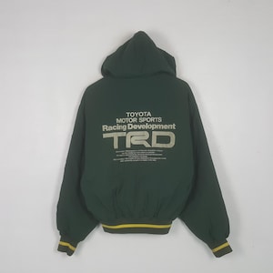 May include: A dark green hooded jacket featuring the text TOYOTA MOTOR SPORTS Racing Development TRD printed in light gray on the back. The jacket has long sleeves with ribbed cuffs and a ribbed hem, both accented with two thin yellow stripes.