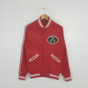 May include: A red varsity jacket featuring white striped trim on the collar, cuffs, and hem. The jacket has a button-front closure, two front pockets, and an oval patch on the chest with the text PRL in a stylized font. Long sleeves have white stripes.