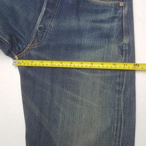 May include: Close-up of blue denim jeans with a measuring tape showing a length of approximately 11 inches. The jeans have a classic five-pocket design and a faded wash, with visible wear and tear.