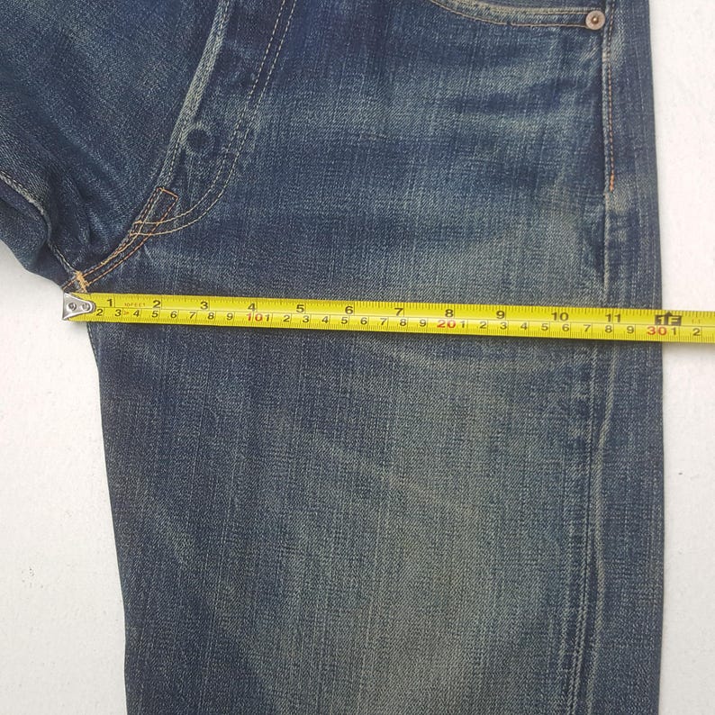May include: Close-up of blue denim jeans with a measuring tape showing a length of approximately 11 inches. The jeans have a classic five-pocket design and a faded wash, with visible wear and tear.
