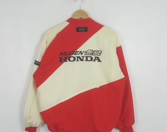 May include: A red and cream-coloured jacket featuring a diagonal colour-blocked design. The back displays the black text MUGEN HONDA with Japanese characters and a small NGK patch near the collar. The jacket has long sleeves and a ribbed hem.