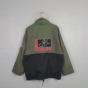 May include: An olive green and black windbreaker jacket featuring a high collar and a rectangular patch on the back. The patch displays the text FUBU in a vertical red box, a white script logo, and the words URBAN GEAR in a red box below.