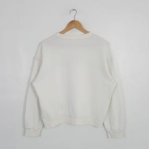 May include: A plain white long-sleeve sweatshirt featuring a crew neckline and ribbed cuffs at the wrists and waist. The garment is displayed on a wooden hanger against a plain white background, showcasing a simple and clean design.