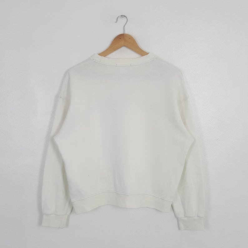 May include: A plain white long-sleeve sweatshirt featuring a crew neckline and ribbed cuffs at the wrists and waist. The garment is displayed on a wooden hanger against a plain white background, showcasing a simple and clean design.