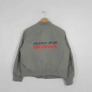 May include: A light gray bomber jacket featuring a ribbed collar, cuffs, and hem. The back displays the text MUGEN in black, Japanese characters for Mugen, and the word HONDA in bold red lettering. The jacket is displayed on a wooden hanger.