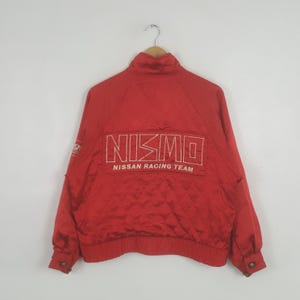 May include: Red Nismo Nissan Racing Team jacket. The jacket has a high collar, long sleeves, and a quilted pattern. The Nismo logo and "Nissan Racing Team" are printed on the back in white.