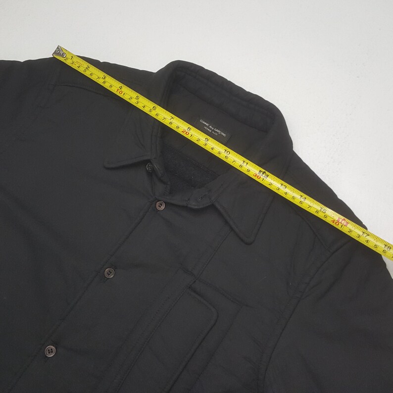 May include: Black button-down jacket with a collared neckline and two front pockets. The jacket has a button closure and a label that reads "COMME des GARCONS". A yellow measuring tape is laid across the jacket.