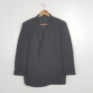 May include: A black blazer with a double-breasted design, hanging on a wooden hanger. The blazer has two rows of buttons and a notched lapel. The garment is made of a solid, dark fabric.