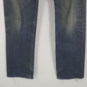 May include: Close-up of the lower portion of a pair of blue denim jeans. The jeans have a faded appearance with distressed areas and frayed edges at the hem. The denim is a medium wash with subtle variations in color.