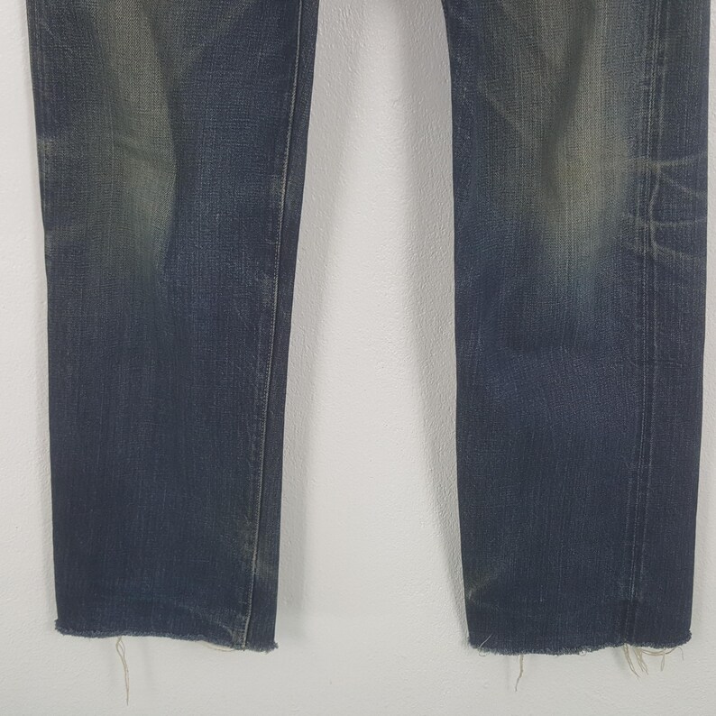 May include: Close-up of the lower portion of a pair of blue denim jeans. The jeans have a faded appearance with distressed areas and frayed edges at the hem. The denim is a medium wash with subtle variations in color.