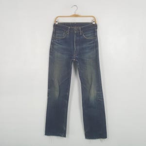 May include: Dark wash denim jeans hanging on a wooden hanger. The jeans have a button fly, classic five-pocket design, and a slightly faded appearance. The bottom hems are frayed.