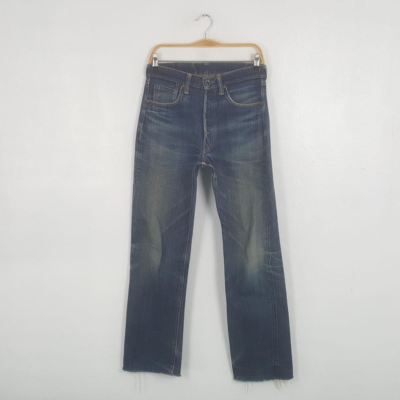 May include: Dark wash denim jeans hanging on a wooden hanger. The jeans have a button fly, classic five-pocket design, and a slightly faded appearance. The bottom hems are frayed.