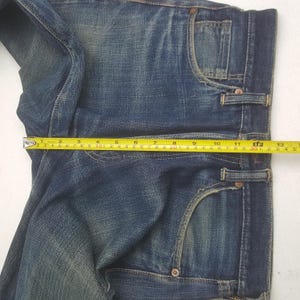 May include: Close-up of blue denim jeans with a measuring tape across the waist. The tape shows a measurement of approximately 13 inches. The jeans have classic five-pocket styling and a faded wash.
