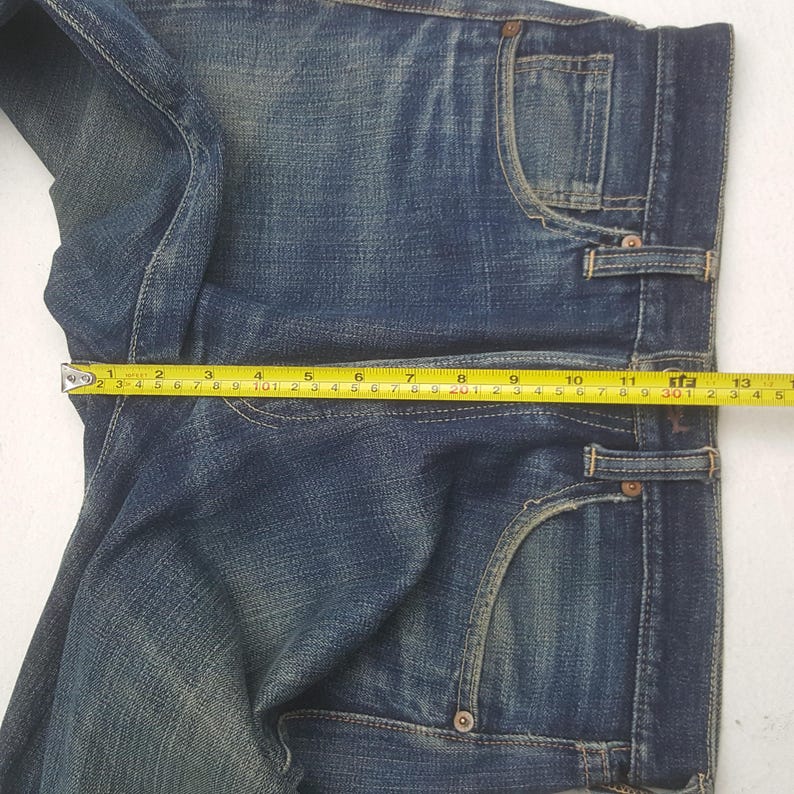 May include: Close-up of blue denim jeans with a measuring tape across the waist. The tape shows a measurement of approximately 13 inches. The jeans have classic five-pocket styling and a faded wash.