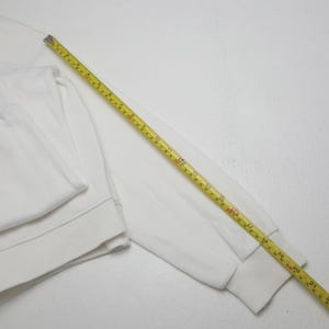 May include: A plain white long-sleeve sweatshirt with ribbed cuffs and hem. A yellow measuring tape is placed diagonally across the sleeve, showing a length of approximately 21 inches from the shoulder seam to the end of the cuff.