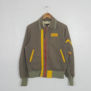 May include: Olive green bomber jacket with a yellow and red stripe down the front. The jacket has yellow accents on the shoulders and pockets, and a small patch on the chest. The cuffs and waistband are a lighter green.