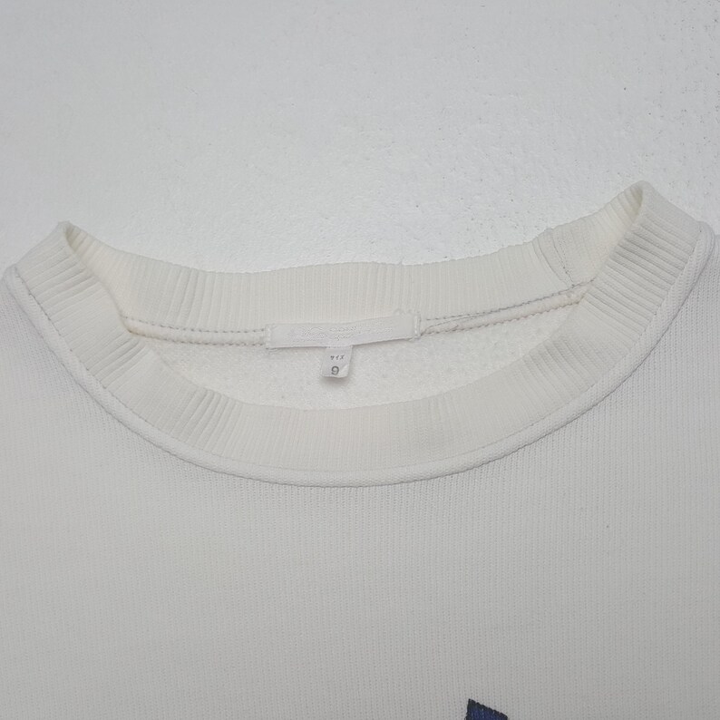 May include: A white crewneck sweatshirt featuring a ribbed collar and a small interior fabric tag at the neckline. The garment is made of a textured material and includes a white label with the number 9 printed in black on the inner collar.