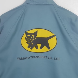 K&ouml;nnte beinhalten: Eine hellblaue Jacke mit einer gelben ovalen Grafik auf der R&uuml;ckseite. Die Grafik zeigt eine schwarze Katze, die ein kleineres schwarzes K&auml;tzchen tr&auml;gt. Unter dem Oval steht der Text YAMATO TRANSPORT CO., LTD. in schwarzen Gro&szlig;buchstaben.
