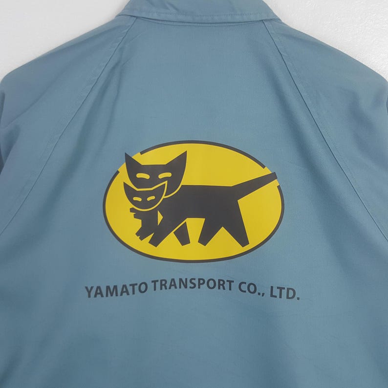 K&ouml;nnte beinhalten: Eine hellblaue Jacke mit einer gelben ovalen Grafik auf der R&uuml;ckseite. Die Grafik zeigt eine schwarze Katze, die ein kleineres schwarzes K&auml;tzchen tr&auml;gt. Unter dem Oval steht der Text YAMATO TRANSPORT CO., LTD. in schwarzen Gro&szlig;buchstaben.