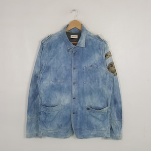 May include: A light blue acid-wash denim jacket featuring a button-down front, two chest pockets, and two lower pockets. The jacket includes shoulder epaulets and a circular camouflage patch on the left sleeve. The interior label reads DIESEL.