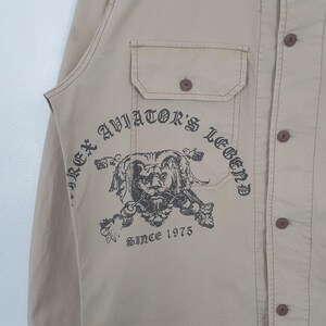 May include: Tan button-up shirt with a chest pocket and a printed graphic of a lion's head with the text "REX AVIATOR'S LEGEND SINCE 1975". The shirt has brown buttons and stitching.
