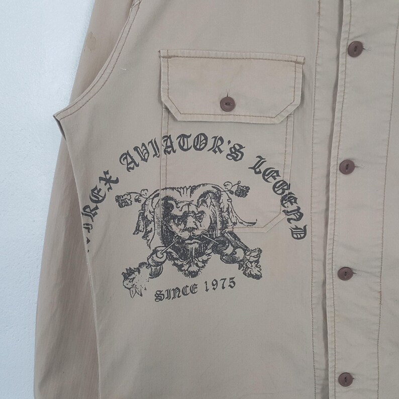 May include: Tan button-up shirt with a chest pocket and a printed graphic of a lion's head with the text "REX AVIATOR'S LEGEND SINCE 1975". The shirt has brown buttons and stitching.