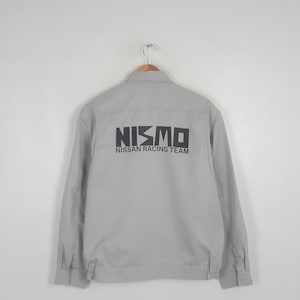 May include: A light gray long-sleeve work jacket featuring a collar, buttoned cuffs, and a black graphic print on the back that reads NISMO NISSAN RACING TEAM in a bold, sans-serif font. The jacket is displayed on a hanger against a plain background.