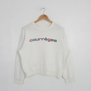 May include: A cream-colored long-sleeve sweatshirt featuring a crew neckline and ribbed cuffs. The word courr&egrave;ges is printed across the chest in lowercase letters, with each letter displayed in a different color including red, green, and blue.